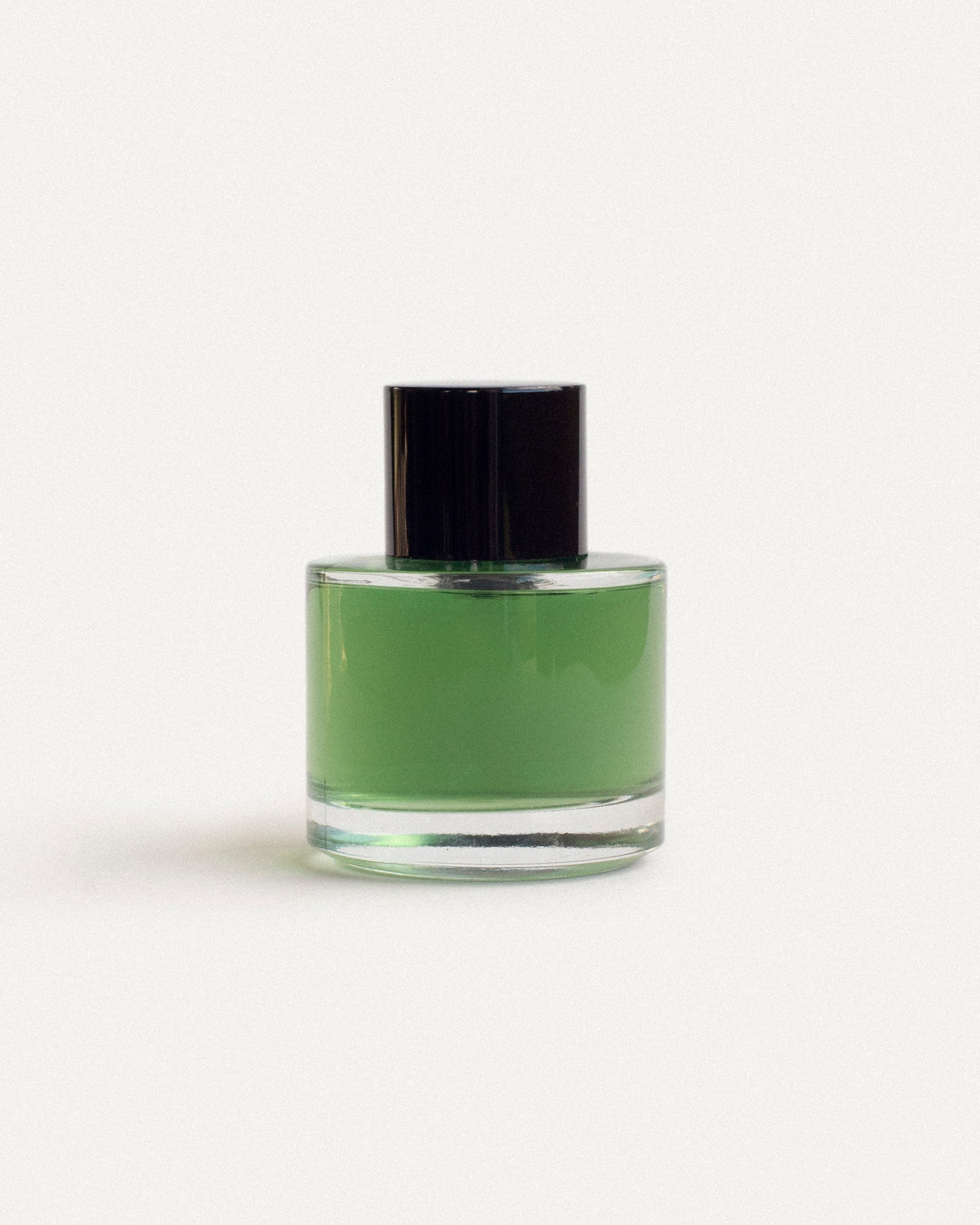 Round clear glass botanical fragrance bottle with matte black cap — vivid emerald green oil, front view on soft off-white — 2 Note Legato Composition, natural perfumery editorial