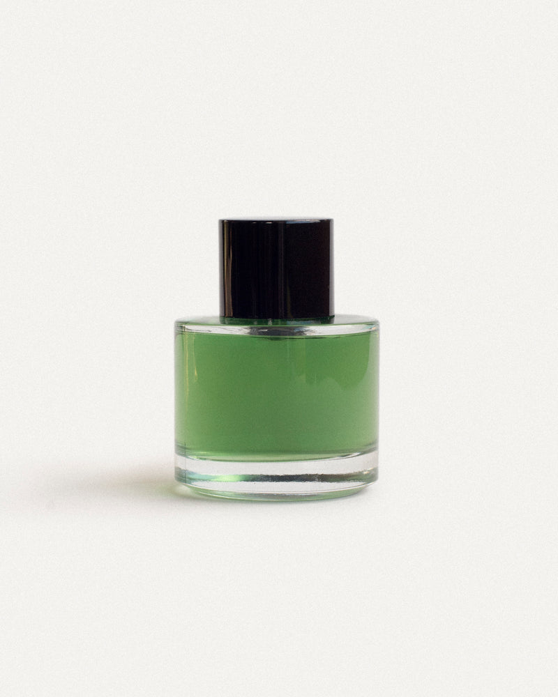 Round clear glass botanical fragrance bottle with matte black cap — vivid emerald green oil, front view on soft off-white — 2 Note Legato Composition, natural perfumery editorial