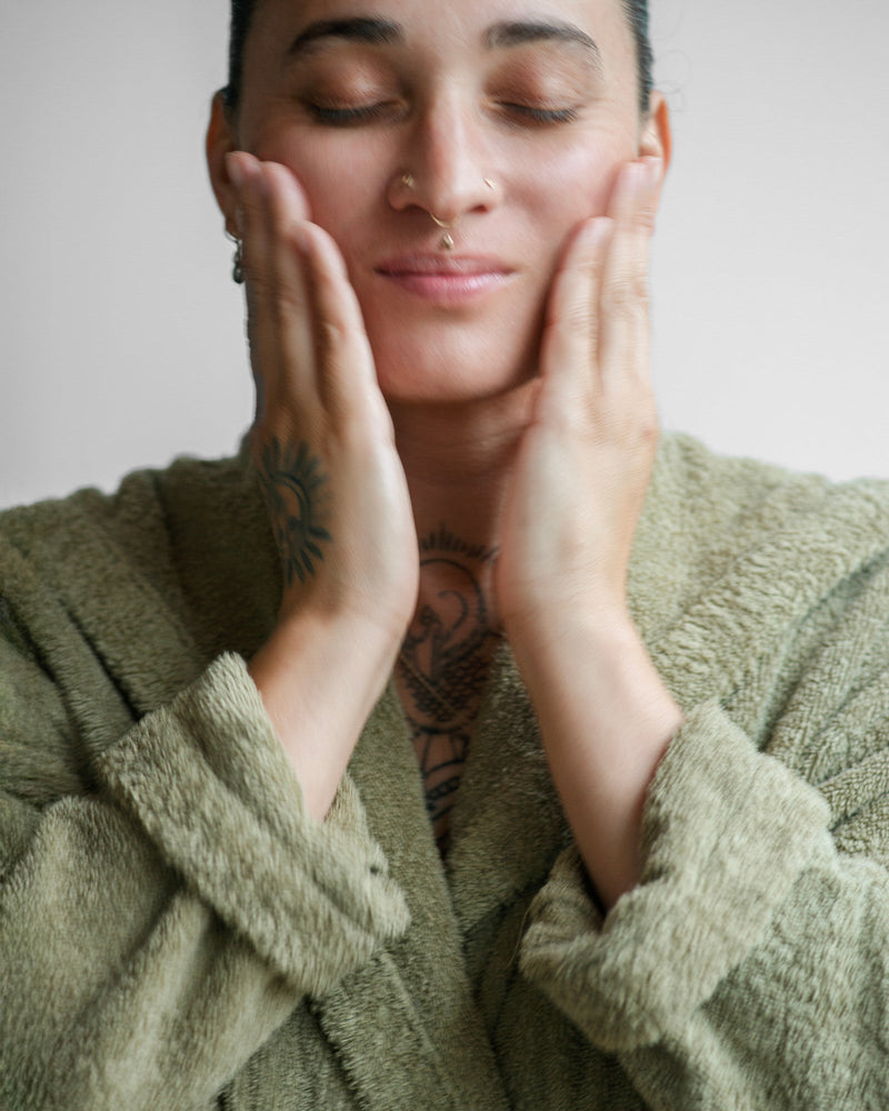Queer nonbinary model with light olive complexion in olive green robe, eyes closed, hands with botanical tattoos pressing cheeks, septum ring visible — botanical natural skincare exfoliation ritual, close-up portrait