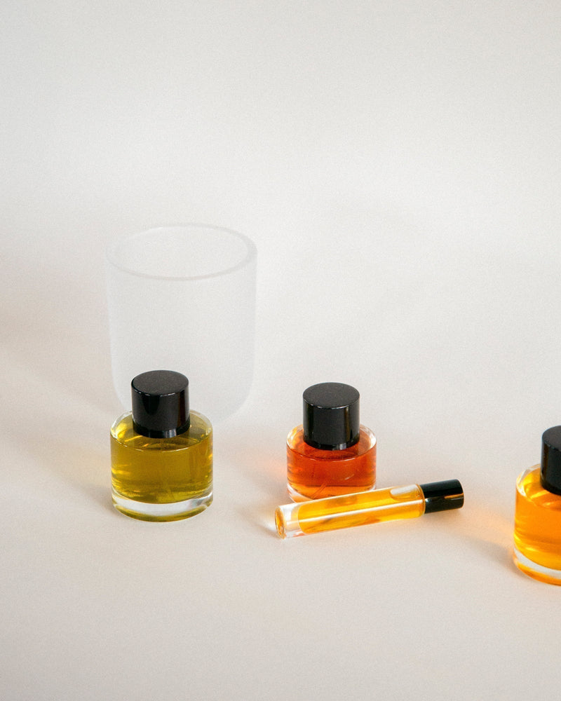 Three 2 Note botanical fragrance bottles in round clear glass with matte black caps — golden yellow, deep orange, and amber natural fragrance oils — with roller bottle and frosted Aromatic Candle vessel on warm off-white surface