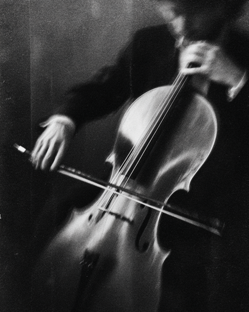 Black-and-white photograph of cellist playing cello with motion blur on bow and strings — dramatic low-key musical editorial for 2 Note Journal