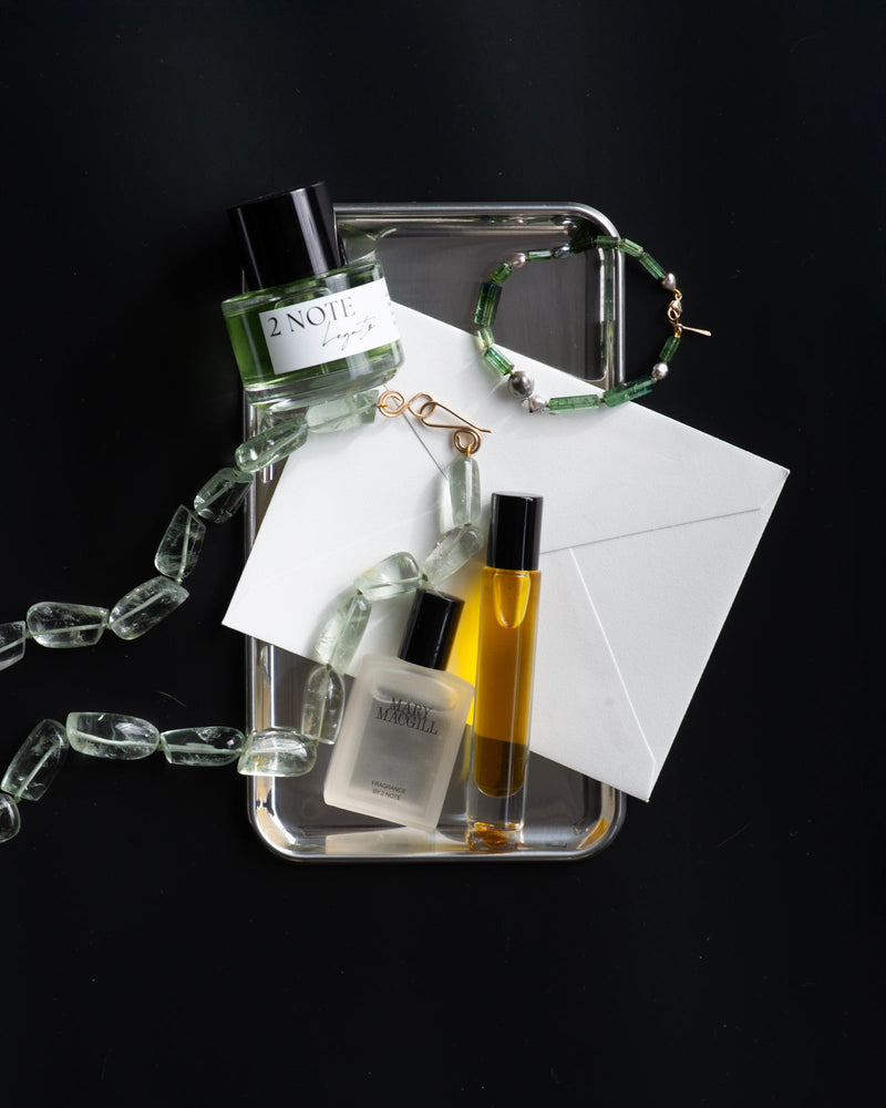 Silver tray on black background with 2 Note Legato fragrance bottle, Mary MacGill x 2 Note collaboration fragrance, amber roller bottle, white envelope, gold wire earrings, and green tourmaline necklace — botanical fragrance collaboration flat-lay