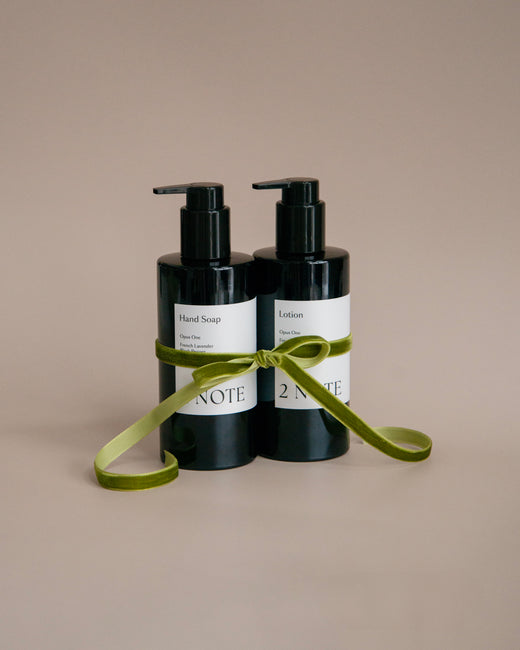2 Note editorial blue set display featuring Botanical Fragrance Chord in a clear glass and black cylindrical Botanical Skincare hand cream and lip balm, styled with a blue plaster wall, black shelf, river stone, and ring.